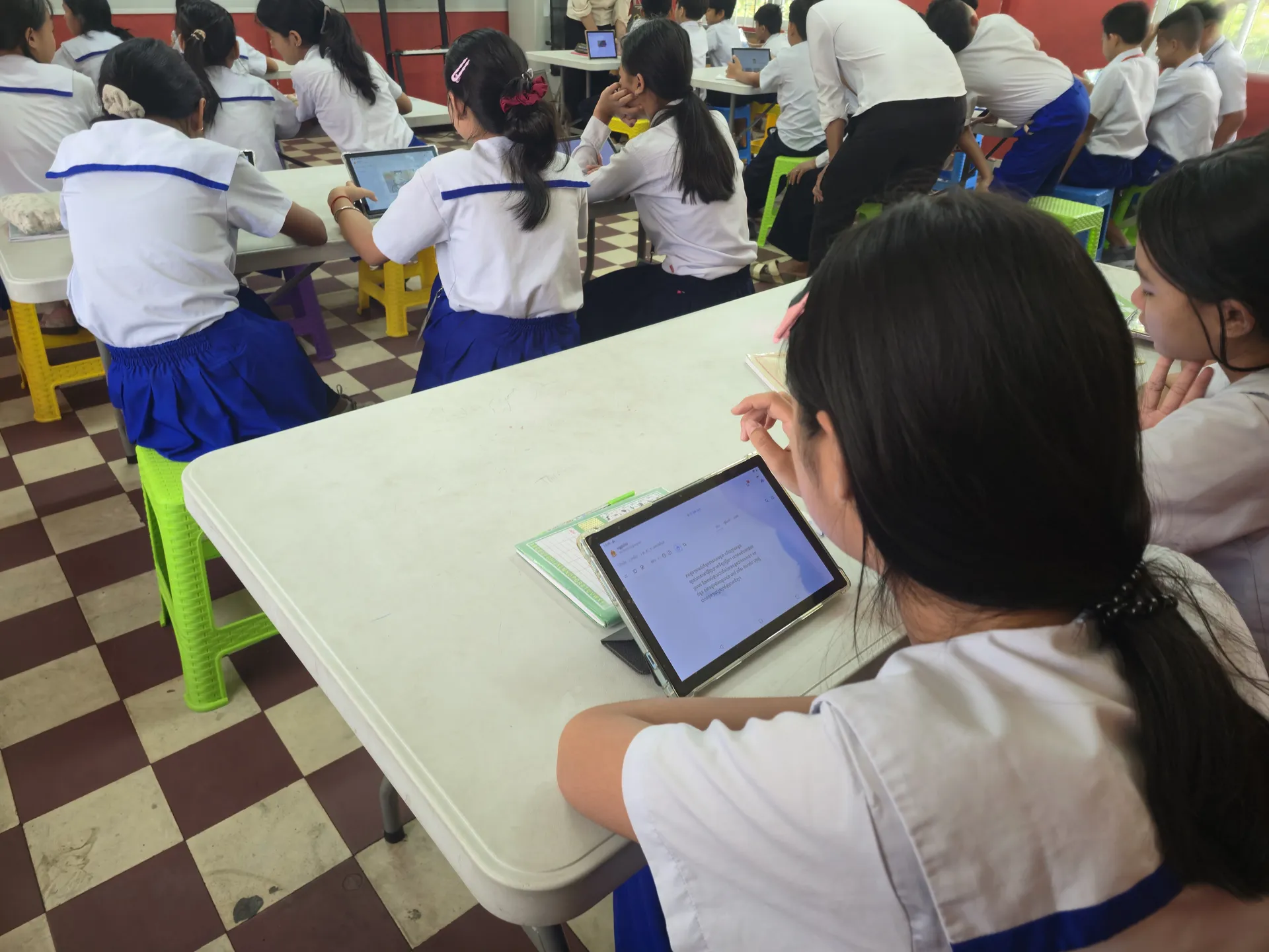 A student reading a page of a book on a tablet in a full classroom.