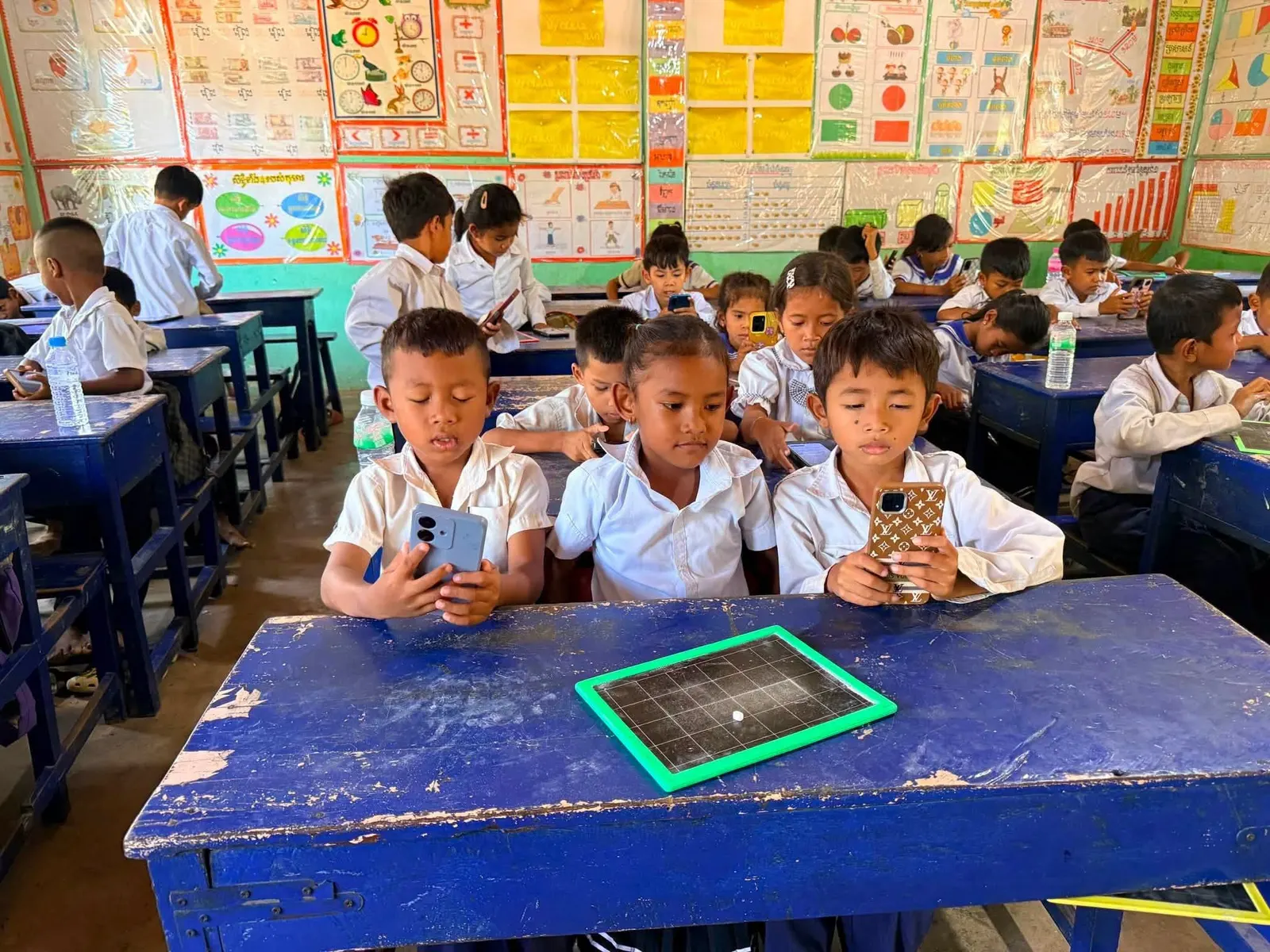 Primary school students at blue wooden desks using the platform on phones, in a classroom with curriculum posters covering every wall.