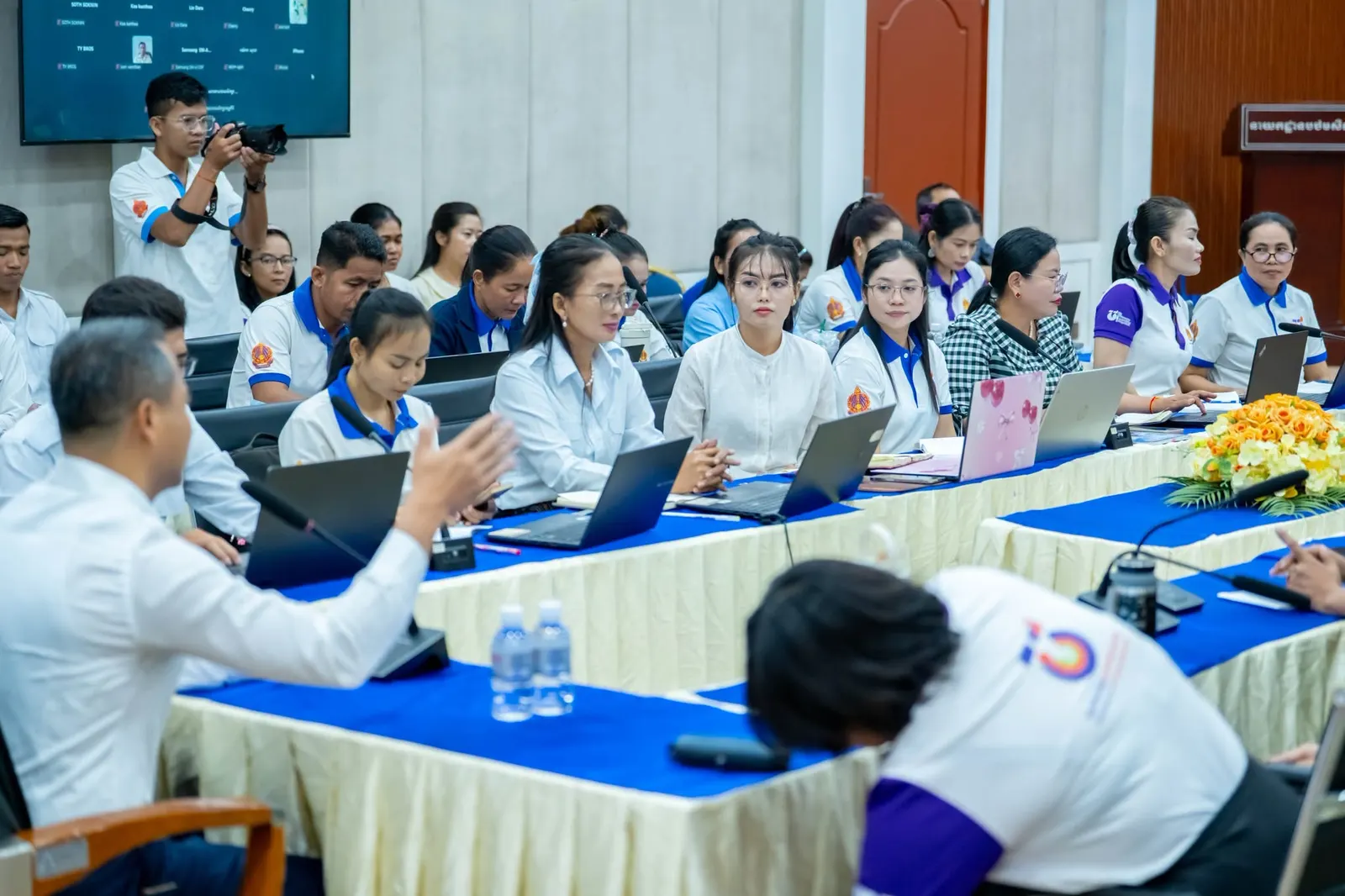Ministry staff and teachers working on laptops around conference tables, a screen at the front listing connected devices.