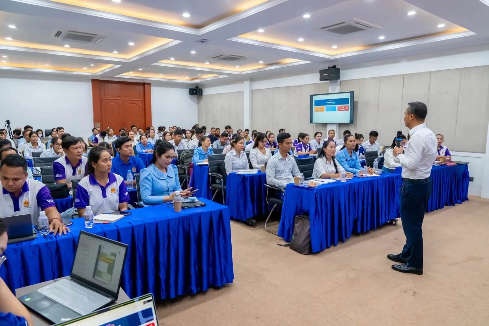 A hall of around a hundred teachers seated at long tables while a presenter demonstrates the platform on a screen behind him.